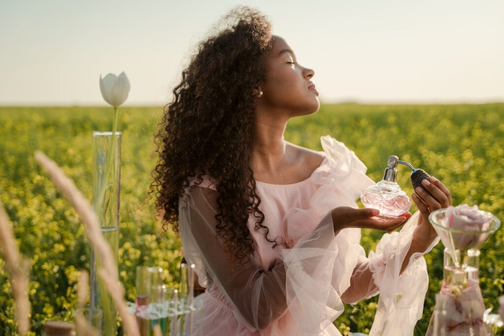 A serene woman in a field wearing a pink dress holds a perfume bottle with eyes closed.